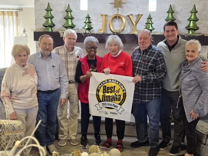 Group of smiling older adults posing together in a holiday-decorated room, holding up a small award banner