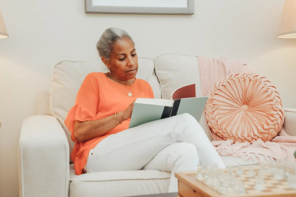 Older woman in an orange top reading a book while relaxing in a white armchair with a pink pillow