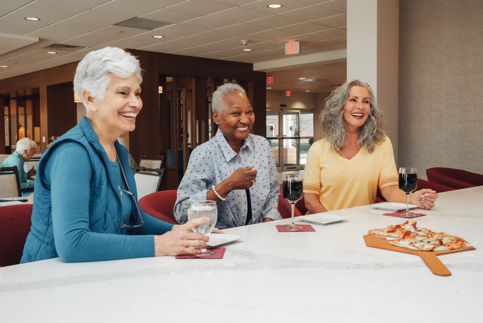 Three smiling older women chatting at a table with glasses of wine and a board of pizza slices