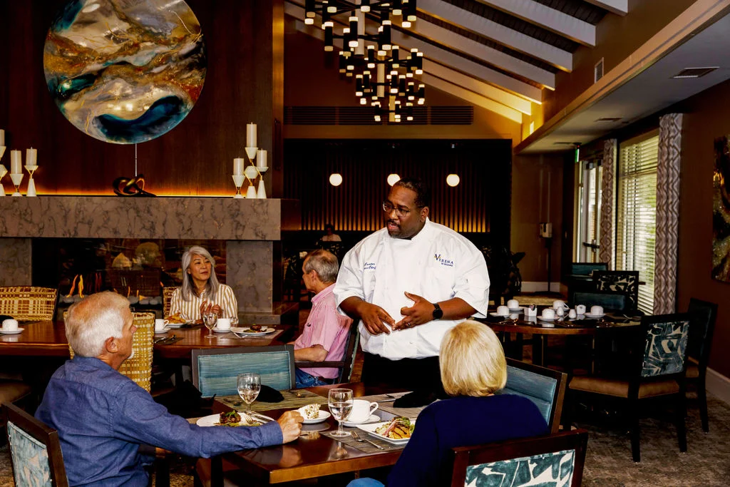 Chef in a white jacket chatting with older residents seated at dining tables in a warmly lit restaurant