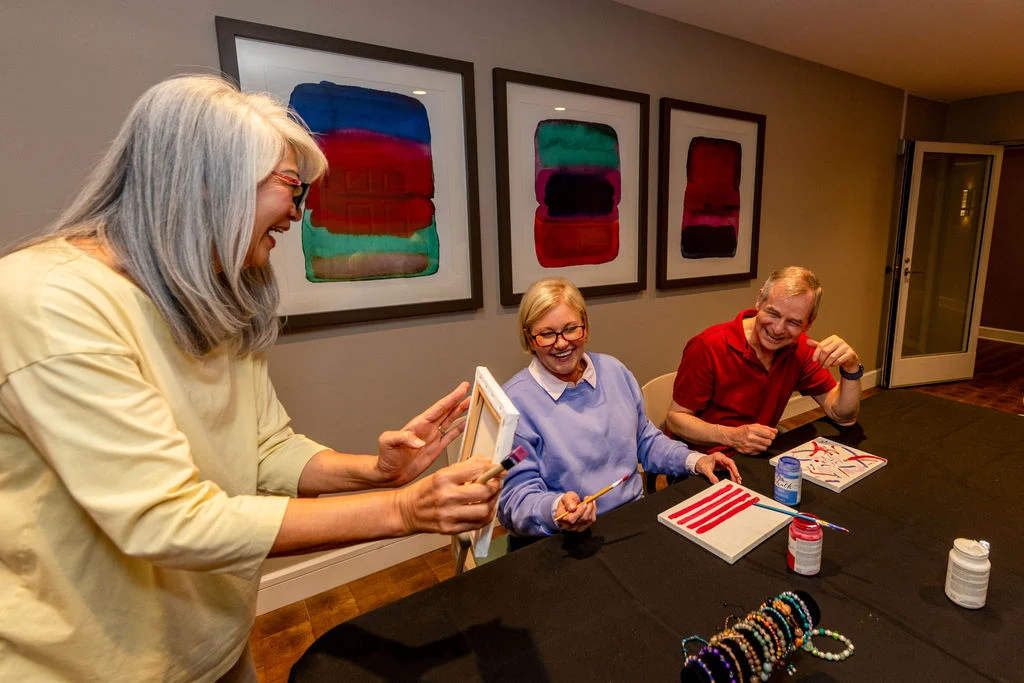 Group of older adults laughing and playing a game around a table, with framed art on the wall behind them