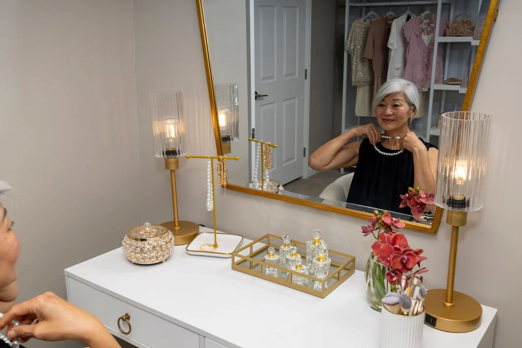 Older woman seated at a white vanity with a gold-framed mirror, lamps and a vase of flowers