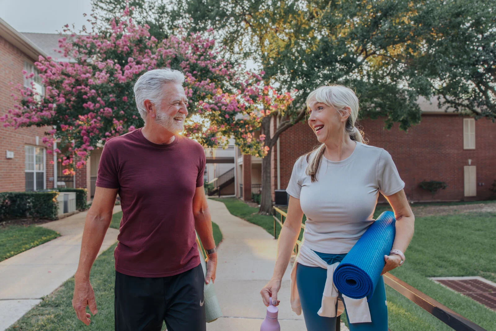 Smiling older couple walking on a path under a flowering tree, the woman carrying a rolled-up yoga mat