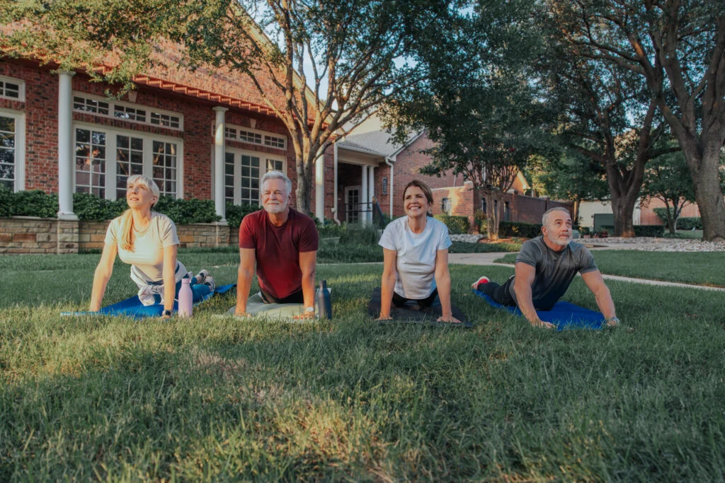 Four older adults doing yoga on mats on a lawn in front of brick buildings