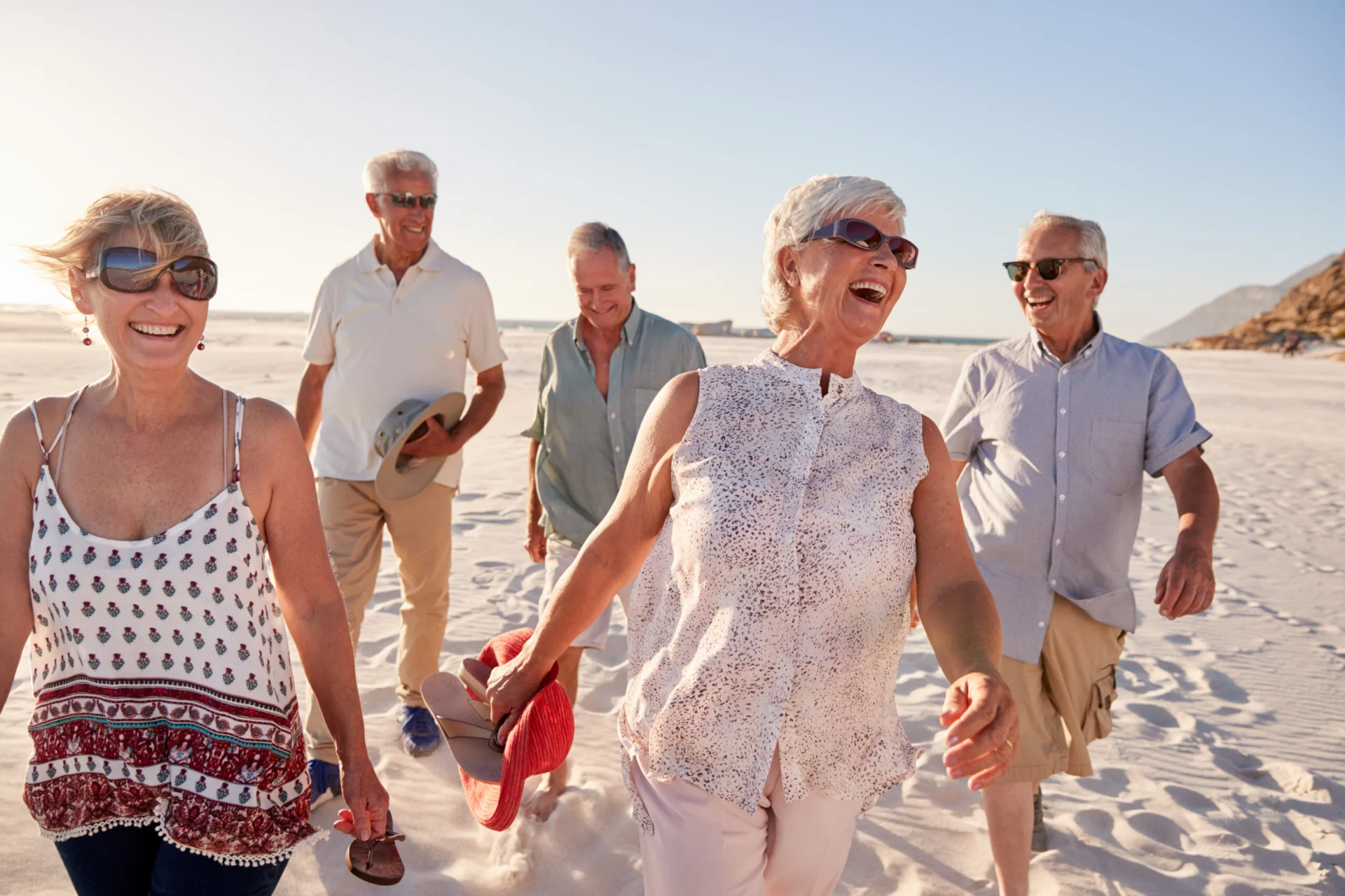 Group of laughing older adults in casual summer clothes walking together on a sandy beach at sunset