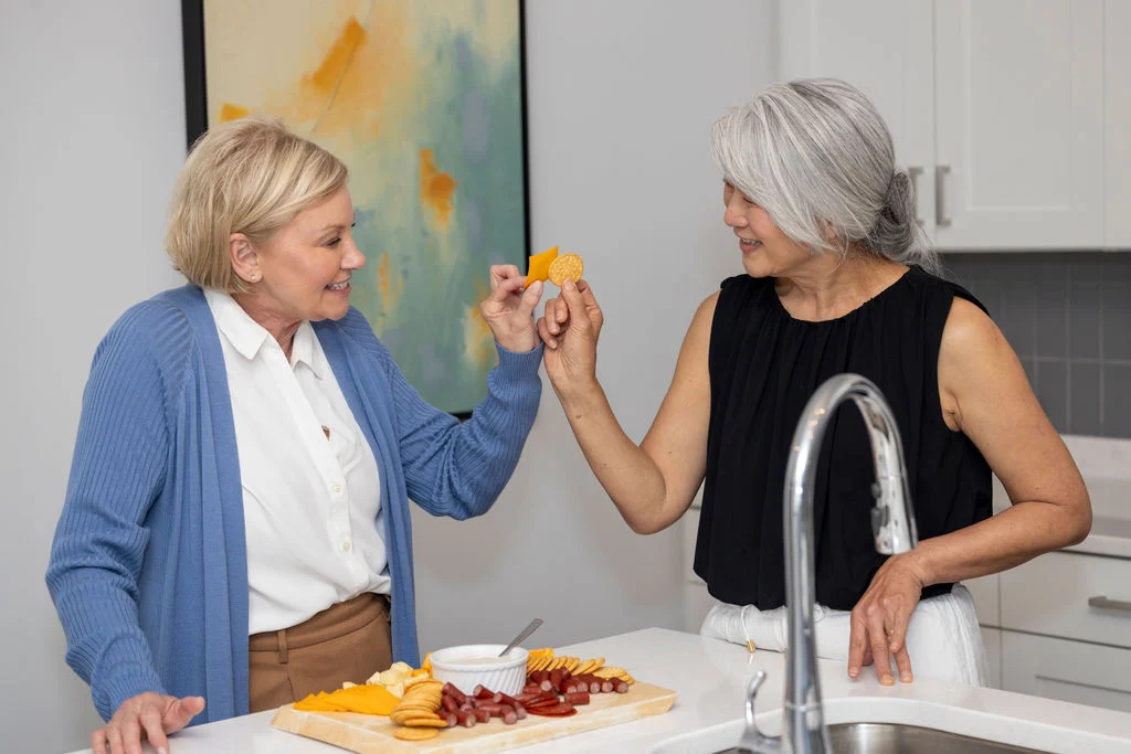 Two older women smiling and raising their hands in a cheerful gesture in a bright kitchen, with a snack board on the counter