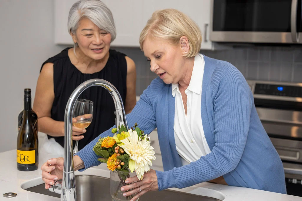 Two older women arranging yellow flowers in a vase at a kitchen sink