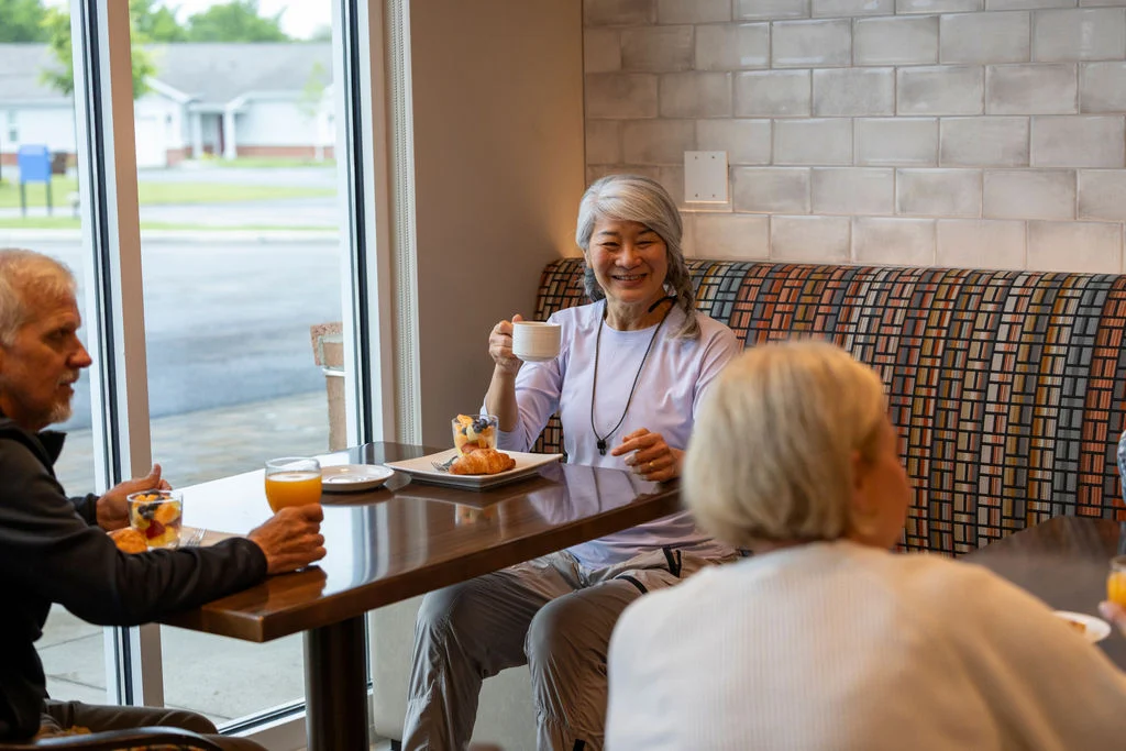 Older adults gathered at a café-style table by a window, sharing drinks and snacks