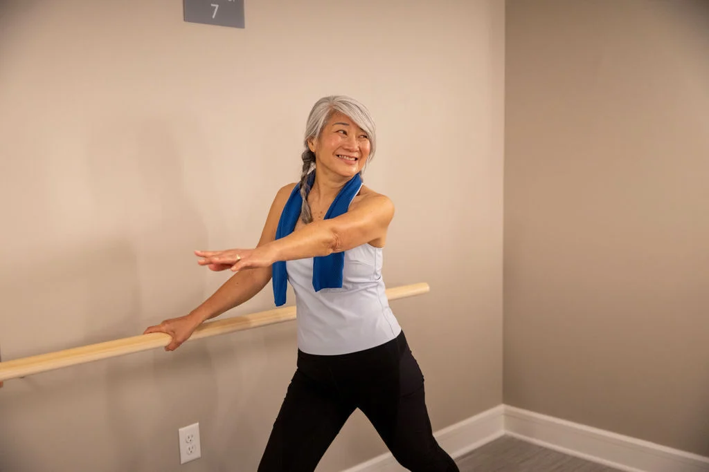 Older woman stretching with one hand on a wall-mounted barre in an exercise studio
