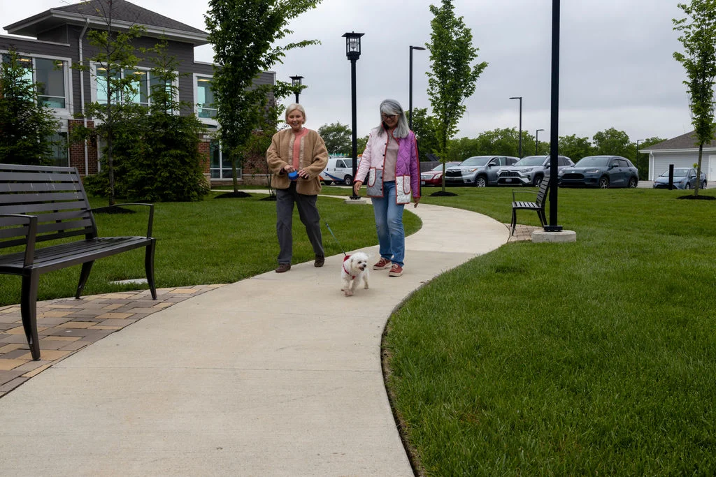 Two people walking a small dog along a curving sidewalk beside a lawn and a bench