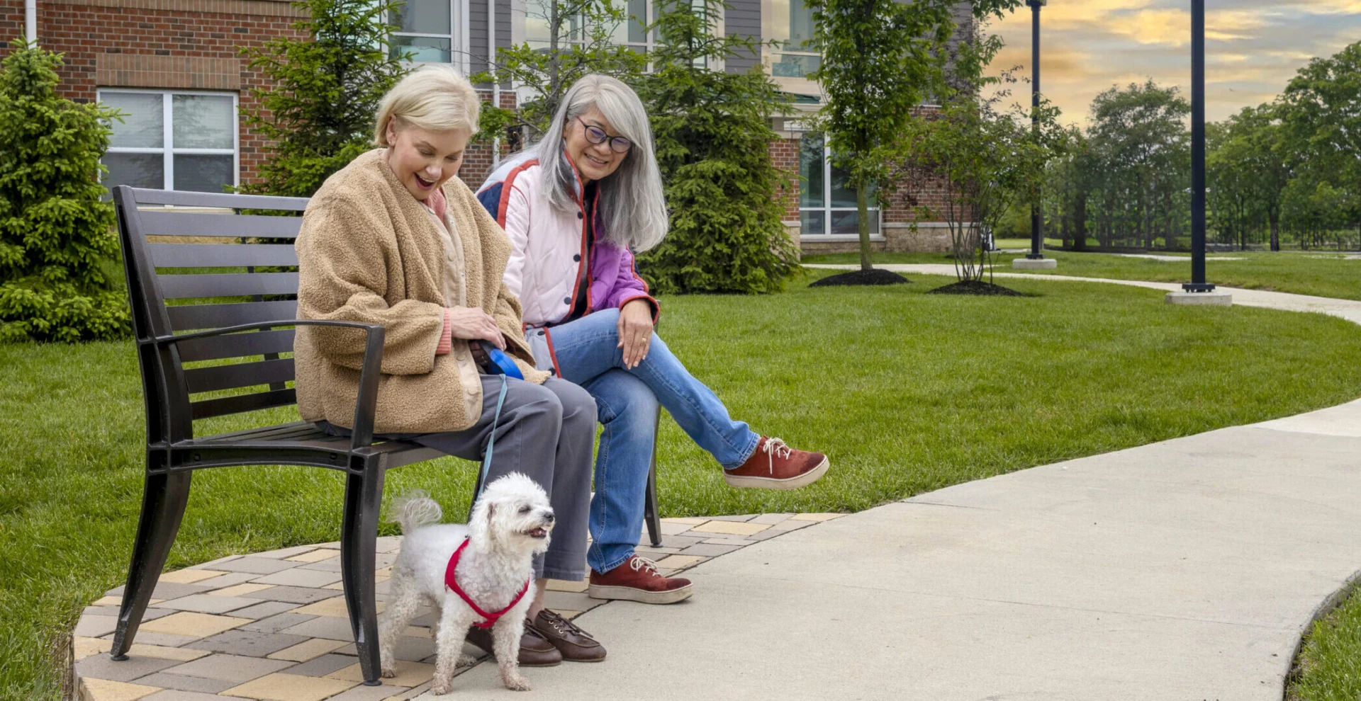 Two older women sitting on a park bench with a small white dog beside them on a paved path through a lawn