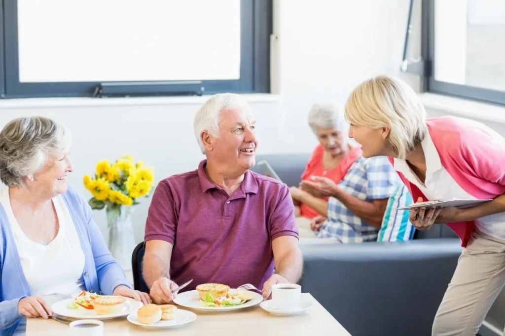 Older adults chatting over a meal at a table with yellow flowers, with a staff member beside them