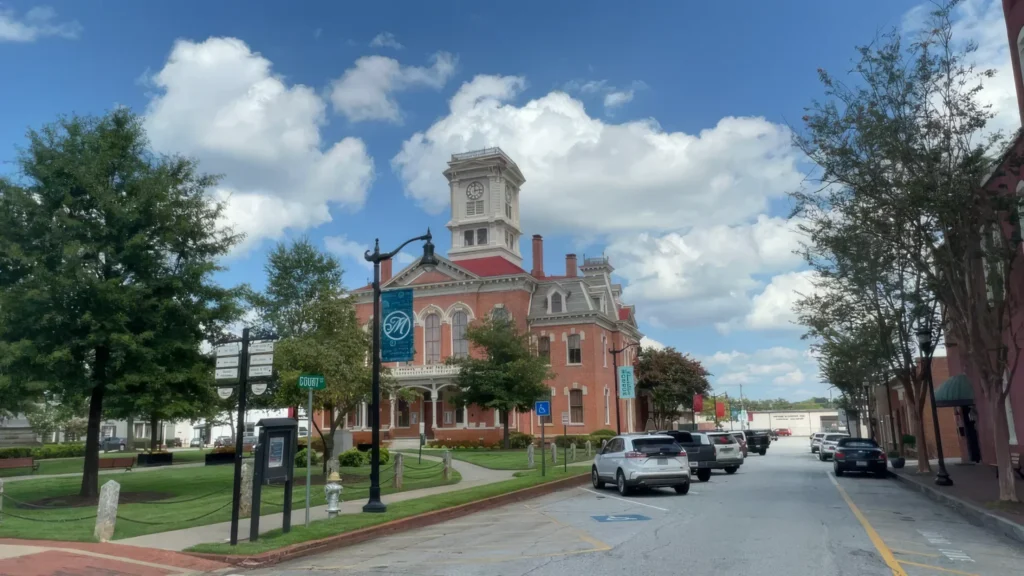 Tree-lined town street with parked cars and a historic brick building topped by a clock tower