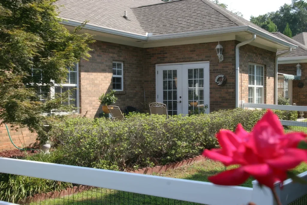 Brick home with French doors and a hedge, with a red flower and white fence in the foreground