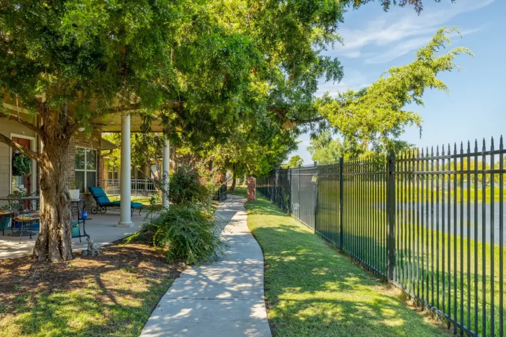 Paved walkway beside a wrought-iron fence and lawn, shaded by large trees