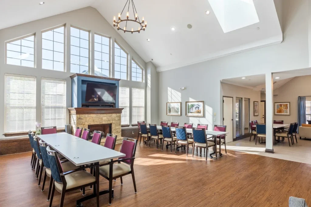 Dining and activity room with long tables, chairs, a stone fireplace with a TV and tall windows under a vaulted ceiling