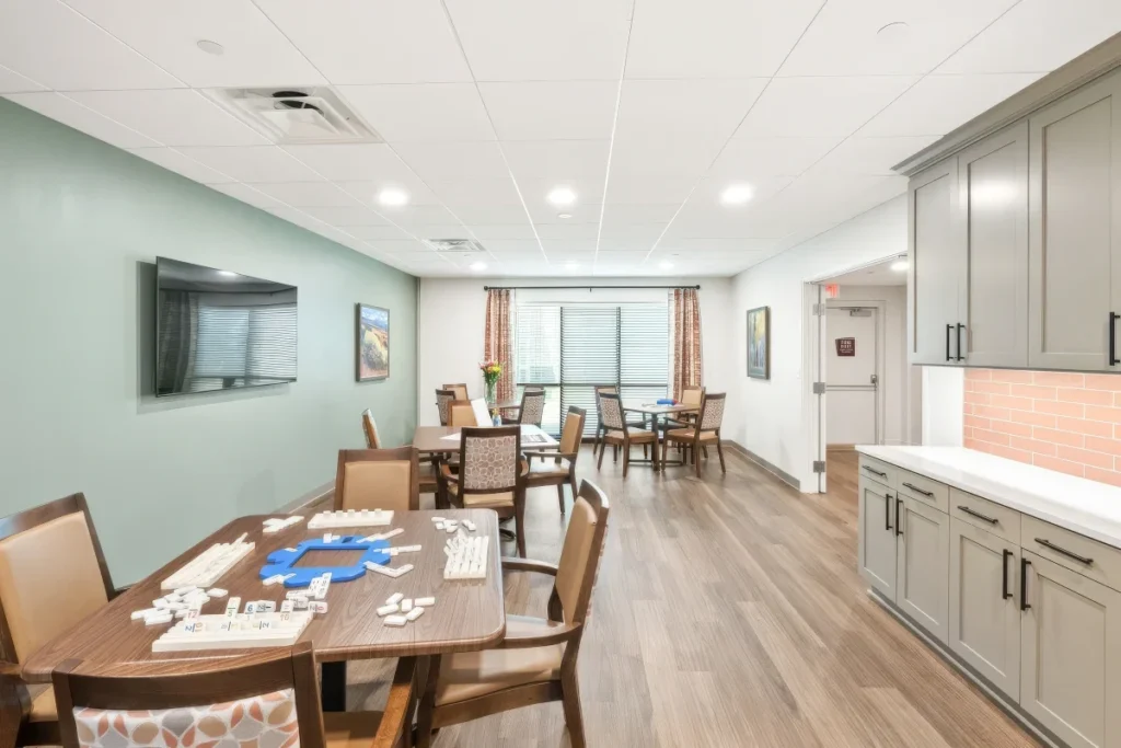 Activity room with a wall-mounted TV, tables set with puzzles and games, and a kitchenette