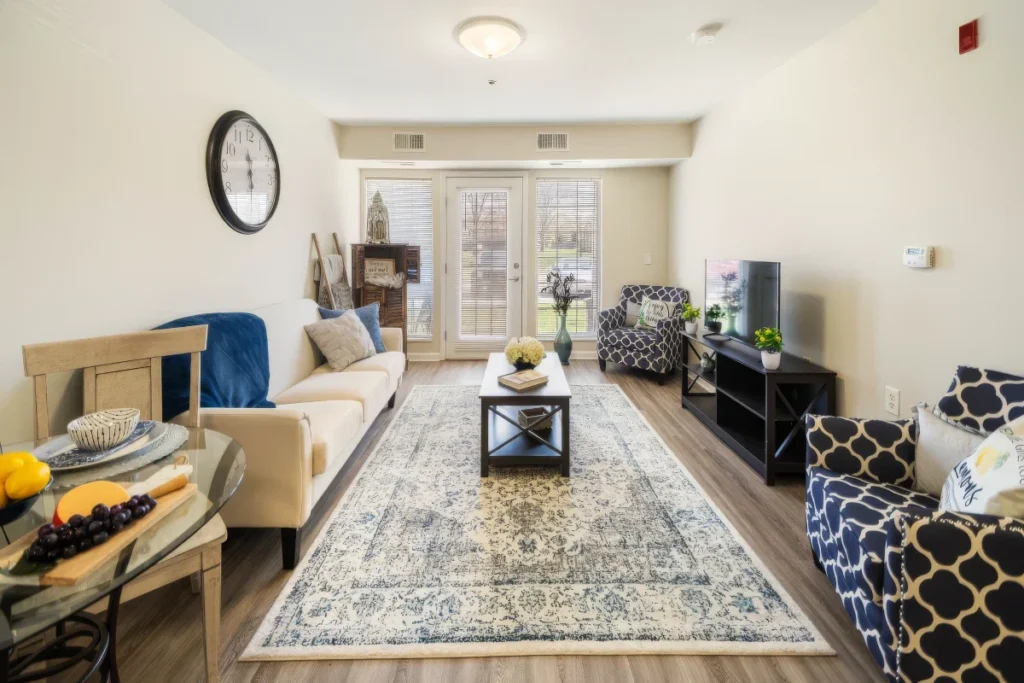Apartment living room with a beige sofa, patterned armchairs, an area rug and French doors, with a dining table at left
