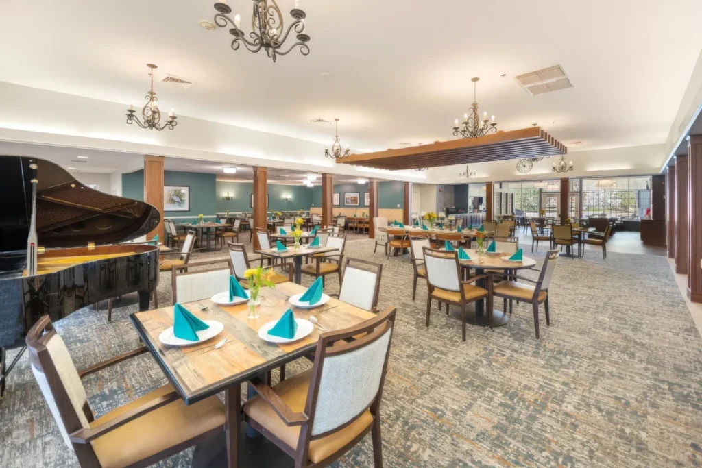Dining room with tables set with teal napkins, cushioned chairs, chandeliers and a piano at the side