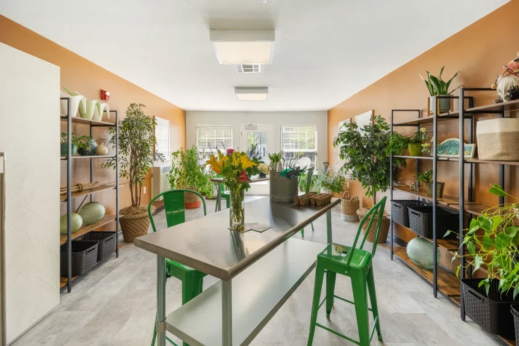 Bright room with shelves of pottery and plants, a long table with a vase of flowers and green stools