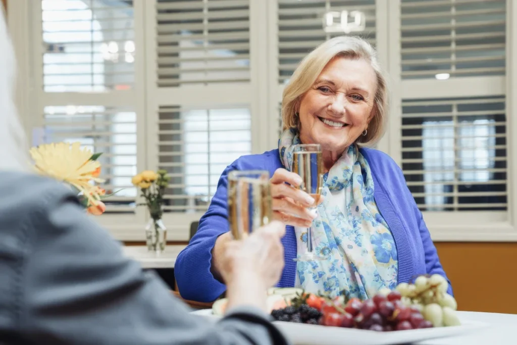 Smiling older woman in a blue cardigan raising a glass at a table with a plate of grapes