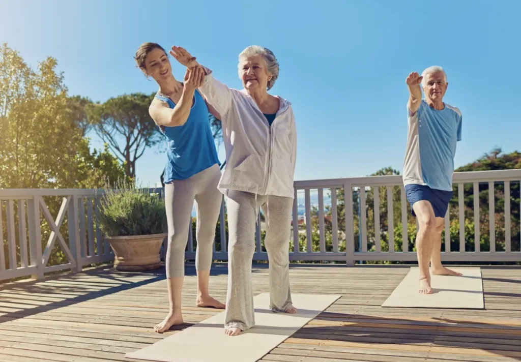 Yoga instructor guiding an older woman's pose while an older man balances on a mat nearby on a sunny wooden deck