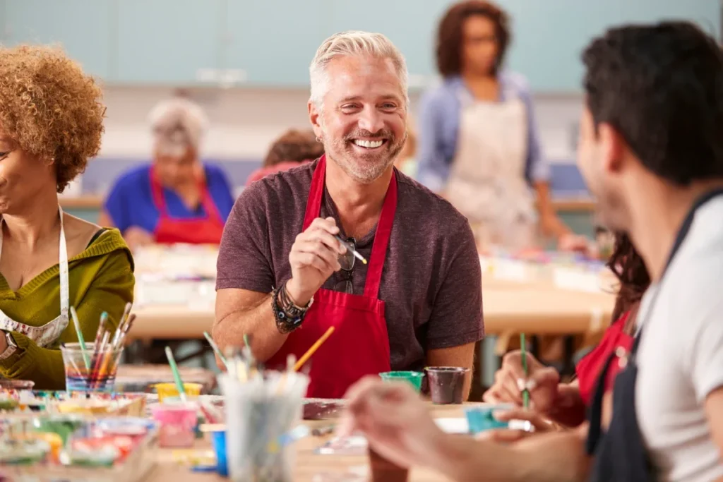 Smiling older man in an apron painting at a table in an art class with others