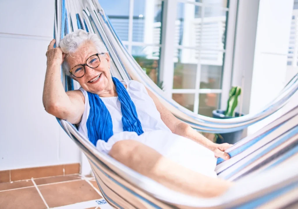 Smiling older woman with glasses relaxing in a striped hammock on a sunny patio