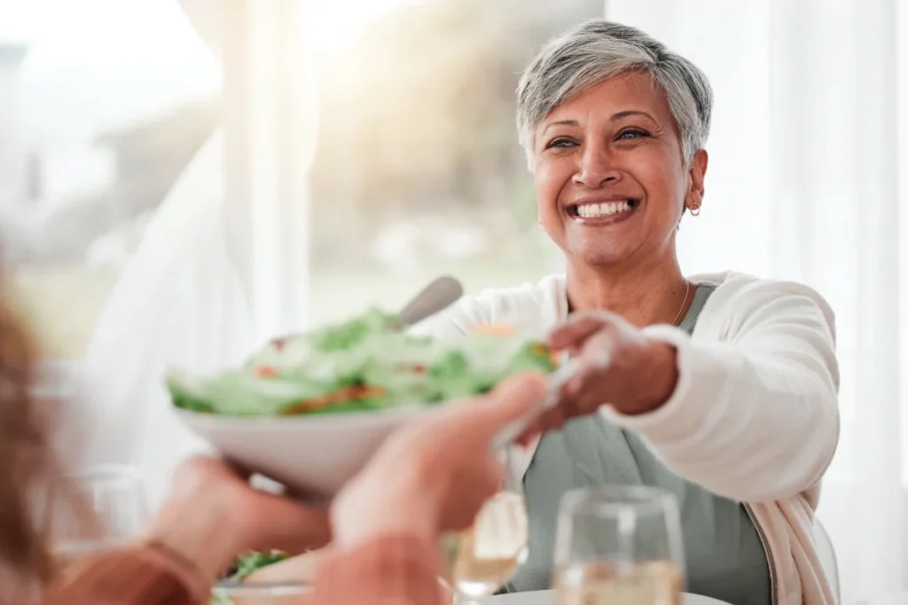 Smiling older woman being handed a bowl of salad at a dining table