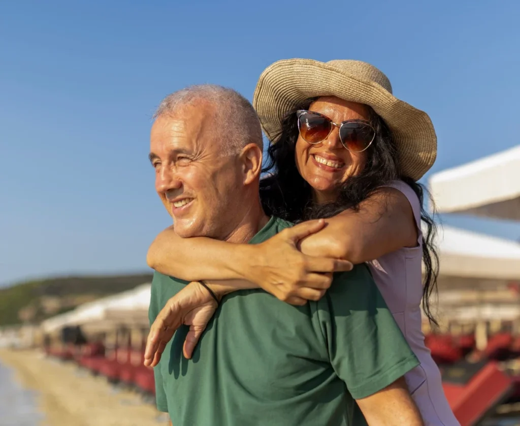 Older woman in a straw hat and sunglasses hugging a smiling older man from behind, with beach loungers in the background