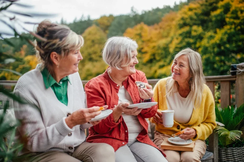 Three older women chatting and sharing snacks on a deck with colorful autumn trees behind them
