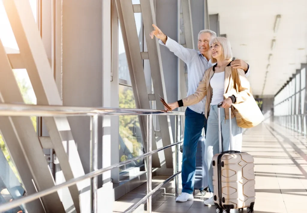 Older couple with rolling luggage walking through a bright airport corridor, one taking a photo
