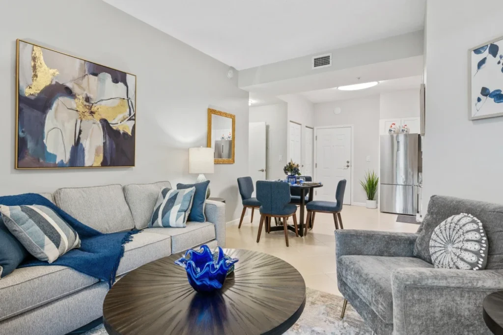Living room with a gray sofa, blue accent pillows and a round coffee table, with a dining table and kitchen beyond