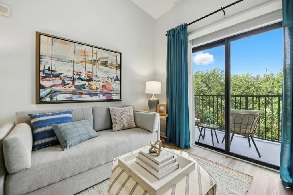 Apartment living room with a gray sofa and a sailboat painting beside a sliding door to a balcony with treetop views