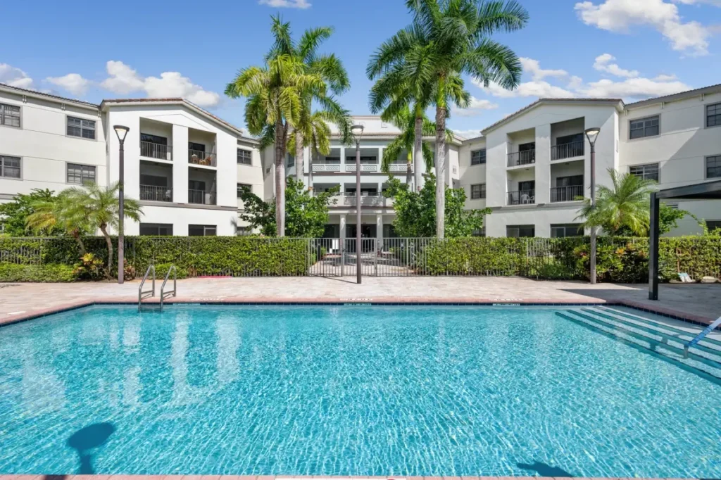 Outdoor swimming pool with palm trees and white apartment buildings behind it under a blue sky