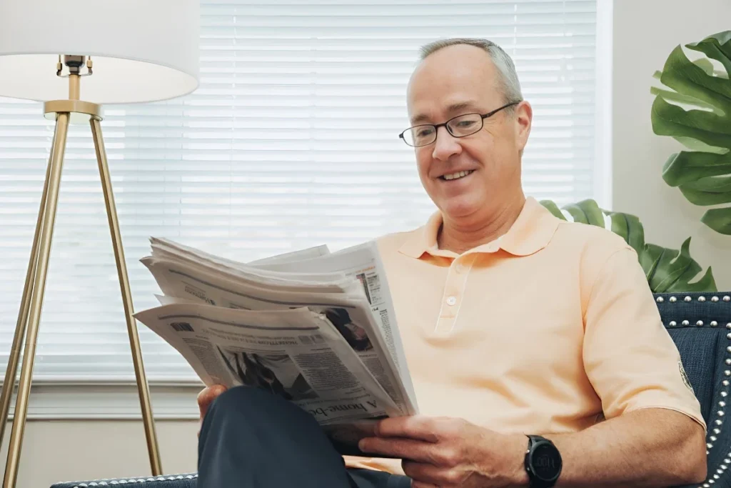 Older man in glasses smiling as he reads a newspaper in an armchair